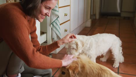 Smiling-caucasian-woman-stroking-her-pet-dogs-eating-from-bowl-in-kitchen-at-home