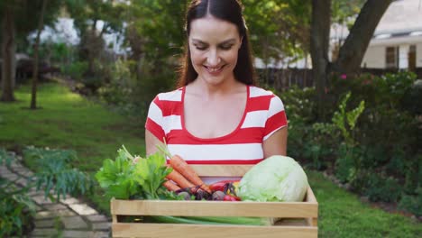 Smiling-caucasian-woman-standing-in-garden-holding-box-of-vegetables