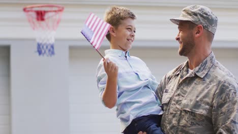 Happy-caucasian-male-soldier-carrying-his-smiling-son,-holding-flags-in-garden-outside-their-house