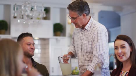 Smiling-caucasian-grandfather-standing-at-table-serving-lemonade-before-family-meal