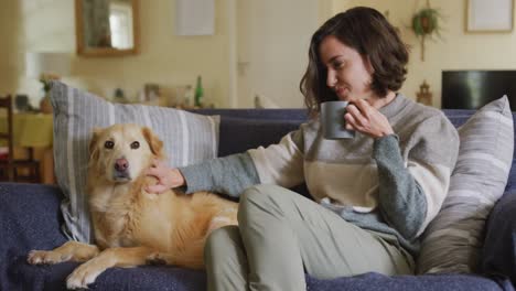 Smiling-caucasian-woman-stroking-her-pet-dog-sitting-on-sofa-at-home