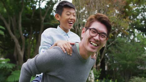 Lächelnder-Asiatischer-Vater,-Der-Seinen-Glücklichen-Sohn-Huckepack-Trägt-Und-Gemeinsam-Spaß-Im-Garten-Hat