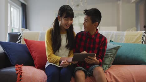 Asian-brother-and-sister-smiling-and-using-tablet-sitting-on-sofa-at-home