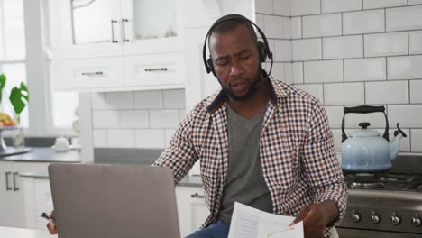 Afroamerikanischer-Mann-Trägt-Ein-Telefon-Headset,-Hält-Ein-Dokument-In-Der-Hand-Und-Spricht-Per-Videoanruf-Auf-Dem-Laptop