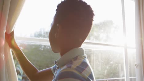 Afroamerikanischer-Junge,-Der-Den-Vorhang-öffnet-Und-Aus-Dem-Sonnigen-Fenster-Schaut,-Das-Von-Der-Sonne-Hinterleuchtet-Wird