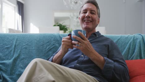 Portrait-of-happy-senior-caucasian-man-sitting-in-living-room-drinking-coffee-and-smiling
