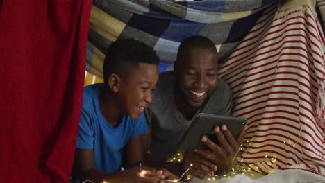 Glücklicher-Afroamerikanischer-Vater-Und-Sohn,-Der-Ein-Tablet-Benutzt-Und-Lachend-Im-Deckenlager-Liegt