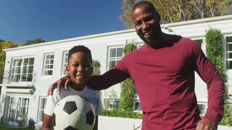 Portrait-of-african-american-dad-and-son-smiling-while-holding-a-football-in-the-garden