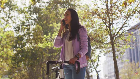Asiatische-Frau-Mit-Fahrrad-Lächelt-Und-Spricht-Auf-Dem-Smartphone,-Während-Sie-Im-Park-Steht