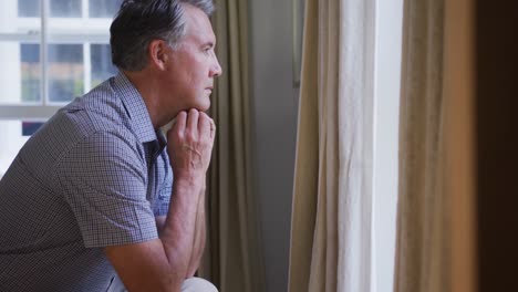 Thoughtful,-sad-senior-caucasian-man-sitting-in-living-room-looking-out-of-window