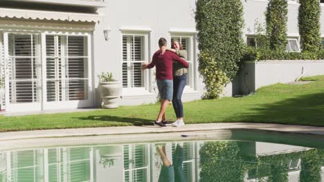 Happy-senior-caucasian-couple-dancing-by-swimming-pool-in-sunny-garden
