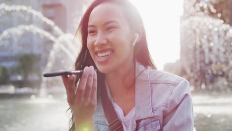Asian-woman-sitting-next-to-fountain-wearing-earphones-and-talking-on-smartphone