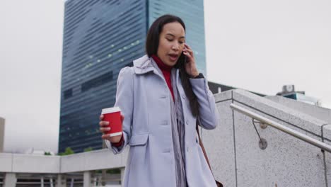Asian-woman-walking-down-the-stairs-talking-on-smartphone-and-holding-takeaway-coffee