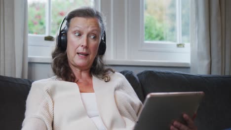 Senior-caucasian-woman-sitting-wearing-headphones-and-talking-during-video-call-using-tablet