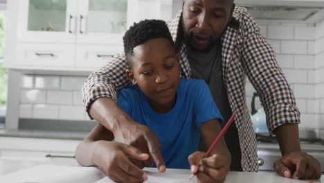 Glücklicher-Afroamerikanischer-Vater,-Der-Sich-über-Seinen-Sohn-Beugt,-Der-Am-Küchentisch-Sitzt-Und-Bei-Seinen-Schularbeiten-Hilft