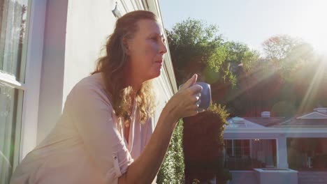 Thoughtful-senior-caucasian-woman-standing-on-sunny-balcony-drinking-coffee-and-looking-around