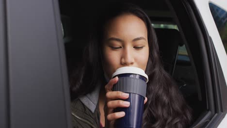 Asian-woman-with-lowered-face-mask-looking-out-of-the-window-and-drinking-coffee-sitting-in-the-car