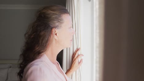 Thoughtful-senior-caucasian-woman-standing-in-sunny-room-looking-out-of-window