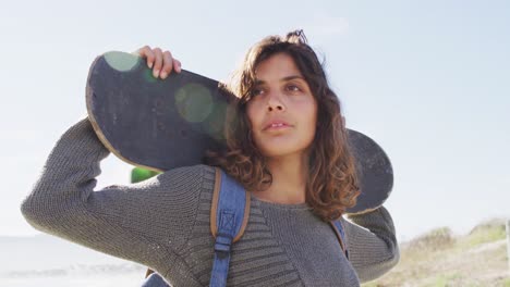 Mixed-race-woman-holding-skateboard-behind-head-standing-in-the-sun-enjoying-the-view-by-the-sea