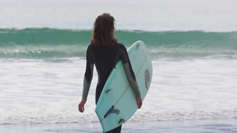 Rear-view-of-mixed-race-woman-walking-into-the-sea-carrying-surfboard