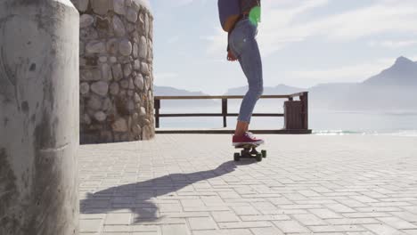 Rear-view-of-mixed-race-woman-skateboarding-on-sunny-promenade-by-the-sea