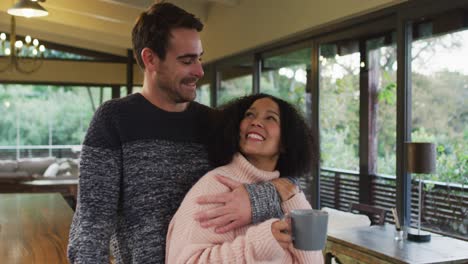 Happy-diverse-couple-talking,-embracing-and-drinking-coffee-in-dining-room