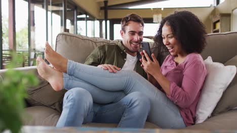 Mixed-race-couple-smiling-while-looking-in-smartphone-together-on-the-couch-at-vacation-home