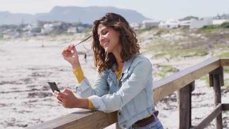 Happy-mixed-race-woman-leaning-on-wall-using-smartphone-and-smiling-on-sunny-promenade-by-the-sea