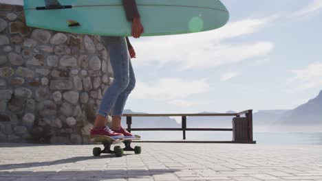 Happy-mixed-race-woman-skateboarding-carrying-surfboard-on-sunny-promenade-by-the-sea