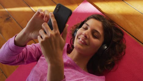 Happy-mixed-race-woman-in-headphones,-lying-on-yoga-mat-using-smartphone-and-smiling-in-sunny-room