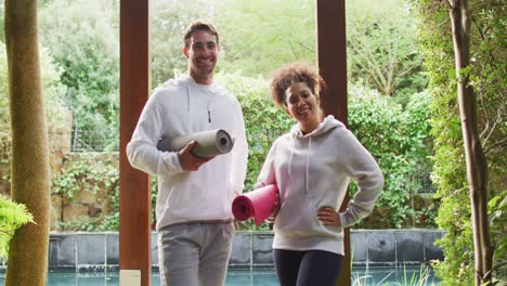 Portrait-of-mixed-race-couple-standing-together-holding-yoga-mats-at-vacation-home
