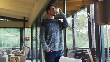 Thoughtful-caucasian-man-looking-out-of-window-and-drinking-coffee-standing-in-dining-room