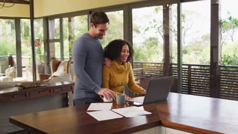 Diverse-couple-talking-in-living-room,-using-laptop,-looking-at-paperwork-and-paying-bills