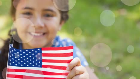 Animation-of-white-circles-moving-over-mixed-race-girl-holding-american-flag