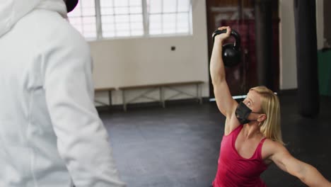 Fit-caucasian-woman-wearing-face-mask-working-out-with-kettle-bells-with-male-trainer-at-the-gym