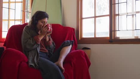 Relaxed-mixed-race-woman-wearing-headphones-sitting-drinking-tea-in-sunny-cottage-living-room