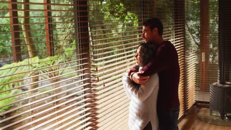 Happy-diverse-couple-embracing-and-smiling-in-living-room,-looking-out-of-window-in-the-countryside