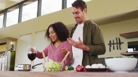 Happy-diverse-couple-preparing-a-meal-together-in-kitchen,-stirring-food-and-laughing