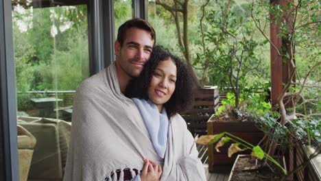 Smiling-mixed-race-couple-wrapped-in-sheets-embracing-each-other-in-the-balcony-at-vacation-home
