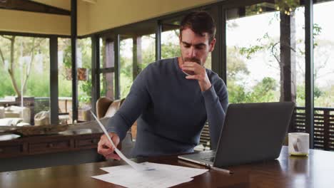 Thoughtful-caucasian-man-reading-documents-and-using-laptop-at-vacation-home