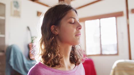 Relaxed-mixed-race-woman-practicing-yoga,-sitting-with-eyes-closed-in-sunny-cottage-bedroom