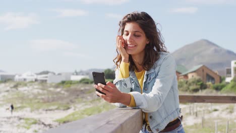 Happy-mixed-race-woman-leaning-on-wall-using-smartphone-and-smiling-on-sunny-promenade-by-the-sea