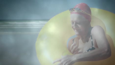 Animation-of-glowing-lights-over-senior-woman-with-yellow-inflatable-ring-by-sea-and-clouds