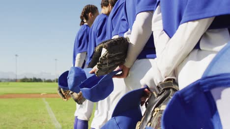 Midsection-of-team-of-female-baseball-players-standing-in-line-on-field-holding-helmets-and-gloves