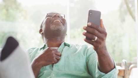 Happy-african-american-senior-man-sitting-with-feet-up-making-video-call-with-smartphone,-laughing