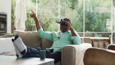 Glücklicher-Afroamerikanischer-älterer-Mann,-Der-Mit-Erhobenen-Füßen-Auf-Der-Couch-Sitzt,-Ein-VR-Headset-Benutzt-Und-Zeigt