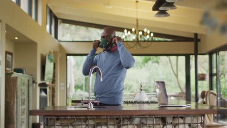 African-american-senior-man-standing-in-kitchen-talking-on-smartphone,-drinking-coffee,-looking-away