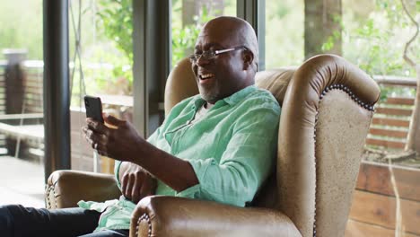 Glücklicher-Afroamerikanischer-älterer-Mann-Im-Wohnzimmer,-Der-Im-Sessel-Sitzt,-Sein-Smartphone-Benutzt-Und-Lacht