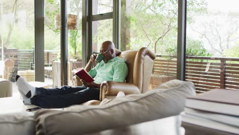 Glücklicher-Afroamerikanischer-älterer-Mann,-Der-Sich-Im-Sessel-Entspannt,-Die-Füße-Hochlegt,-Ein-Buch-Liest-Und-Kaffee-Trinkt