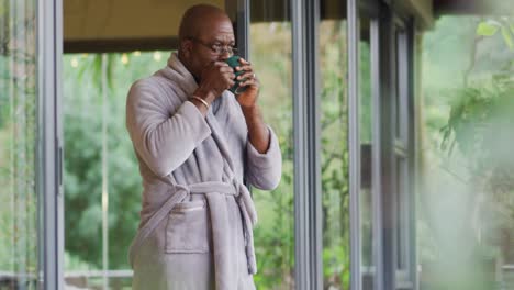 African-american-senior-man-standing-on-balcony-wearing-bathrobe-drinking-coffee-and-enjoying-view